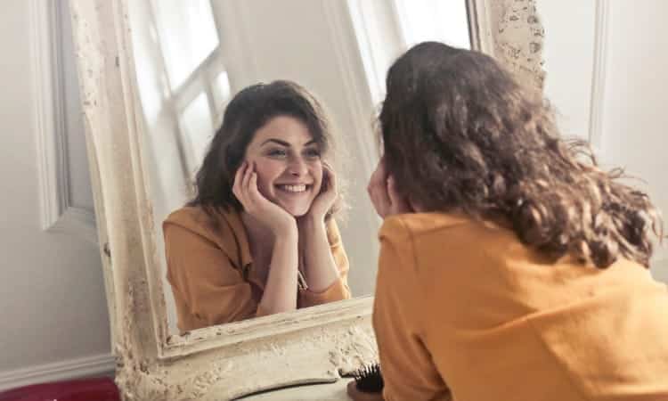 Brunette woman wearing a yellow blouse frames her chin with her hands and smiles into a vintage mirror